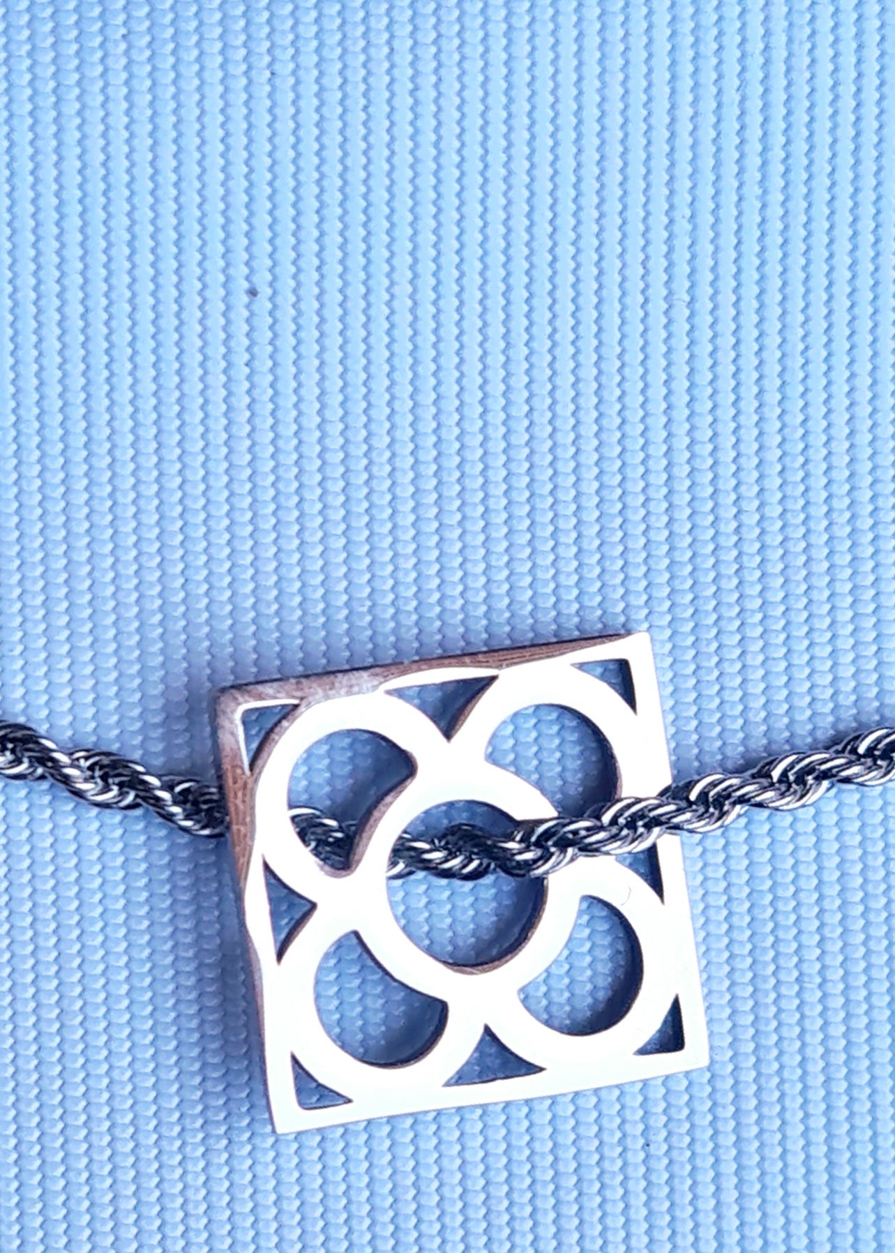 Colgante con la Flor de Barcelona de acero inoxidable cuadrada para mujer. Fondo celeste.