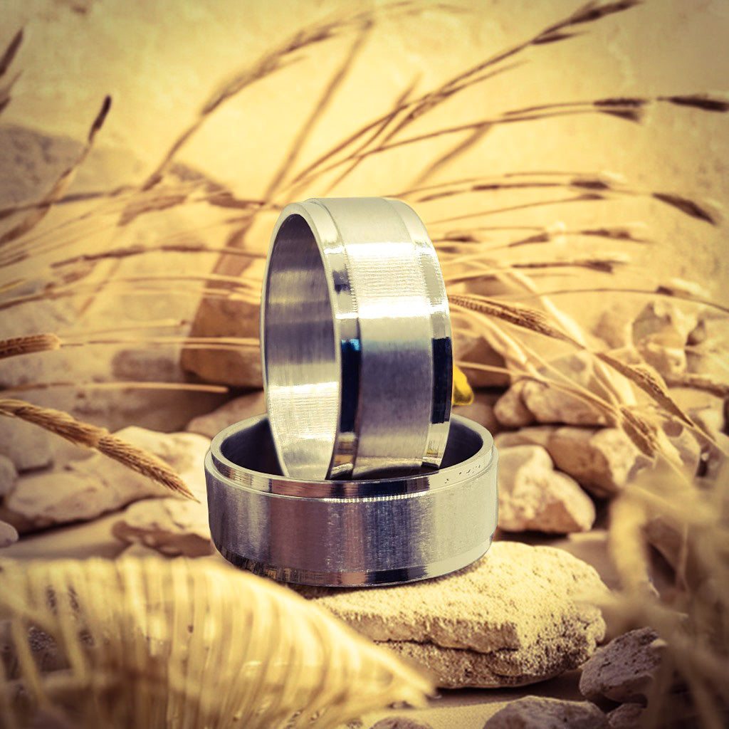 Dos Anillos tipo alianza de matrimonio o pareja en acero inoxidable biselado y ancho de 8mm. Se incluye grabado de nombres y fecha importante gratis. Posa sobre naturaleza.