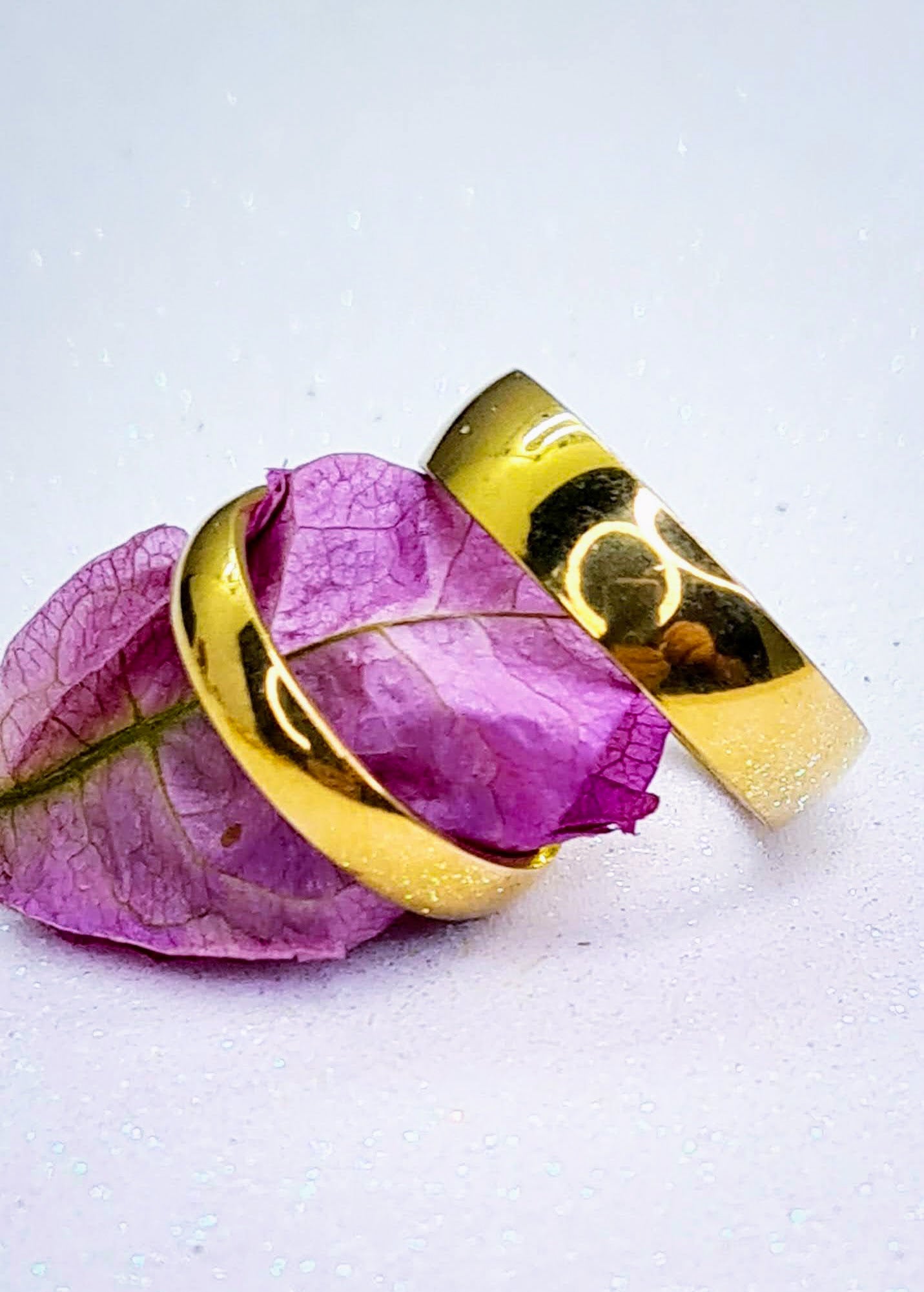 Anillos tipo alianza de matrimonio o pareja en acero inoxidable dorado y ancho de 4mm y 6mm. Se incluye grabado de nombres y fecha importante gratis. Posan sobre fondo blanco brillante con un pétalo de flor.