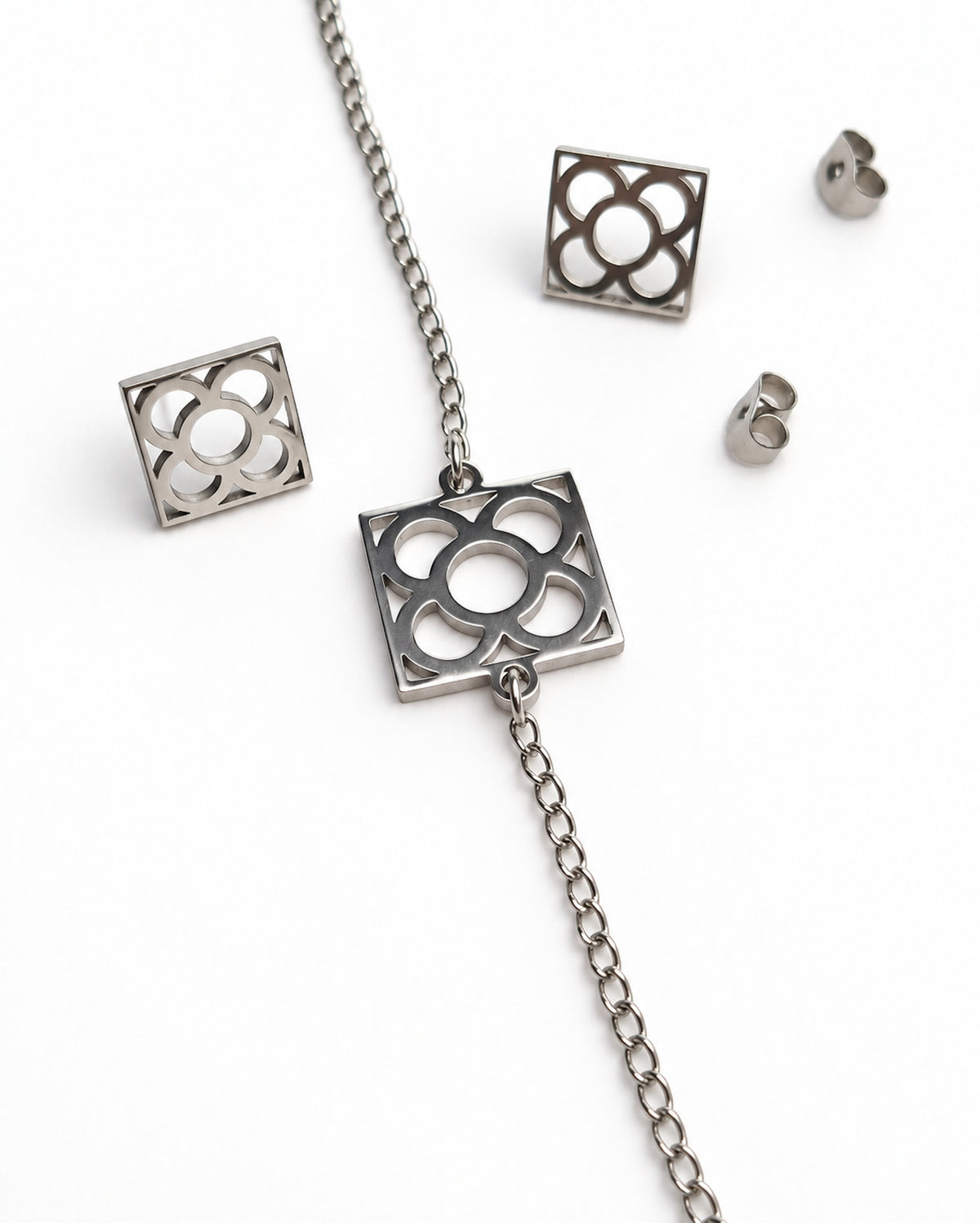 Conjunto plateado Flor de Barcelona con pulsera de cadena y pendientes de tope cuadrados sobre fondo blanco.