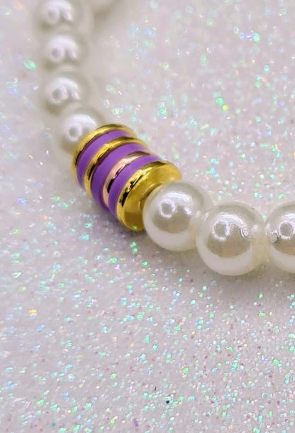 Detalle de pulsera de perlas blancas con discos de cerámica morados y detalles dorados.