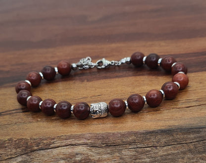 Pulsera unisex de cuentas de sándalo marrón con pieza central de Buda y separadores plateados sobre madera.
